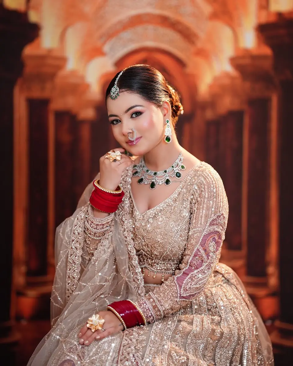 A bride in a champagne and gold lehenga seated with one hand raised to her face, warm lamplit arches behind her.