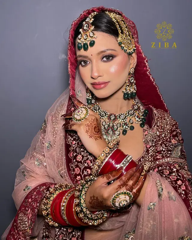 A bride in a maroon and dusty-pink lehenga with an emerald and gold necklace, maang tikka and red chooda, mehndi on both hands.