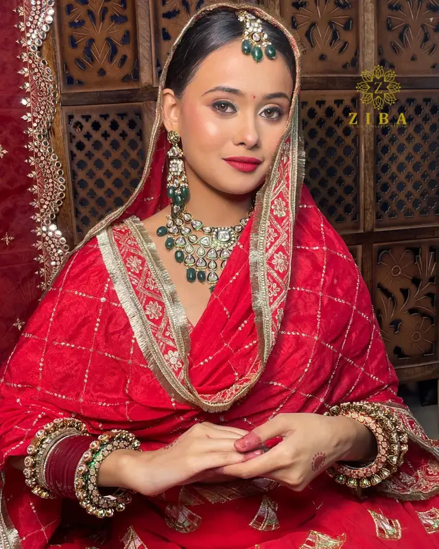 A bride in a red lehenga and dupatta with a green kundan necklace and matching earrings, seated in front of a carved wooden screen.