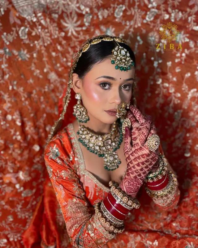 A bride in orange velvet with a wide kundan collar and nose ring, both mehndi-covered hands raised beside her face.