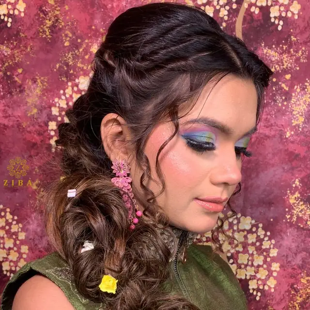 A side view of a woman with blue and green eye makeup and a braided hairstyle threaded with small pink flowers.