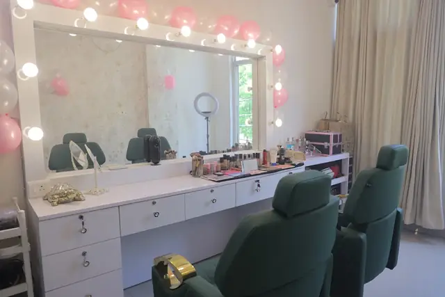 The makeup studio: a long white vanity with a bulb-lit mirror, product trays and two green salon chairs.